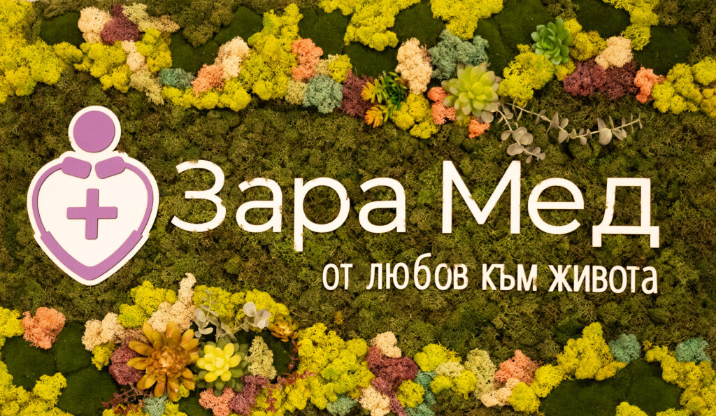 Colorful moss wall featuring a purple medical shield logo with a cross and the Bulgarian text 'Зоя Мед' and slogan below.