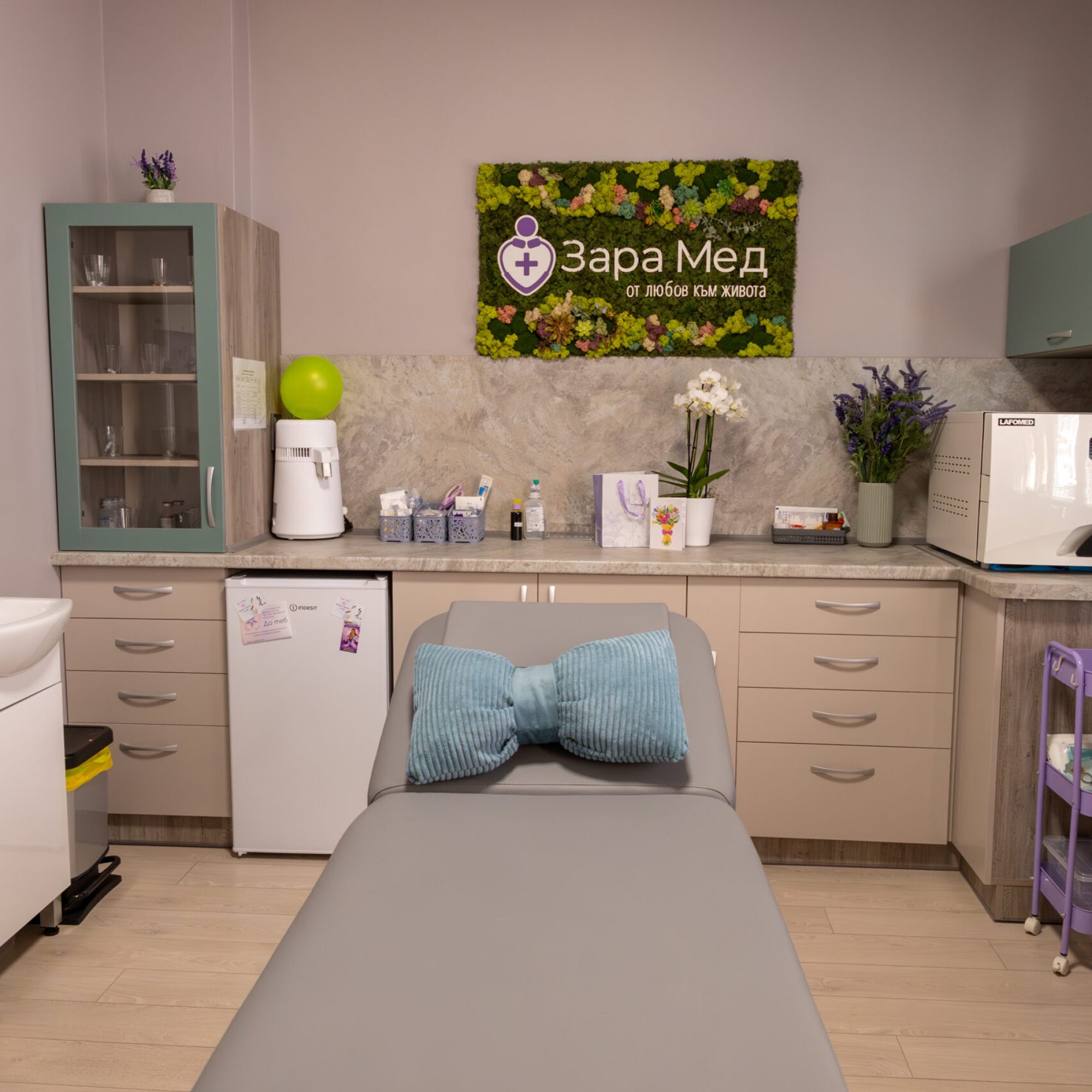 Treatment room with a padded gray exam bed in the foreground and a mossy wall sign behind reading the clinic name in Cyrillic.