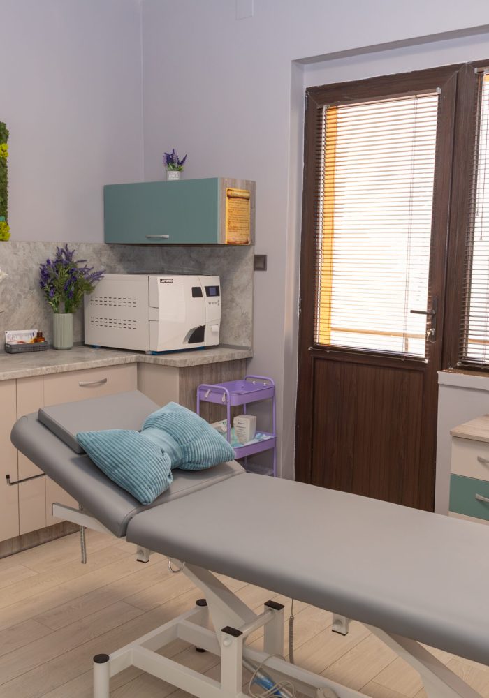 Medical treatment room with a padded examination table and purple pillows, counter with supplies, and a moss wall sign reading 'Зара Мед' on the left. Large windows with blinds on the right.