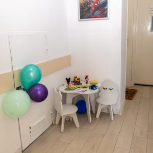 Children's play corner with small table and chairs, balloons, and toy cars on a light wood floor near a white wall and door.
